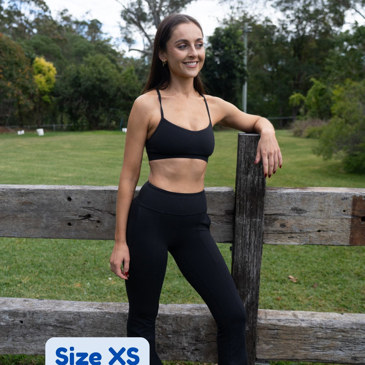 A woman wearing a black sports bra and black high-waisted leggings stands outdoors leaning against a wooden fence, with a grassy park and trees in the background. A white label in the bottom left corner reads 'Size XS'.