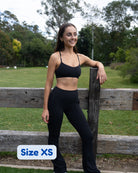 A woman wearing a black sports bra and black high-waisted leggings stands outdoors leaning against a wooden fence, with a grassy park and trees in the background. A white label in the bottom left corner reads 'Size XS'.